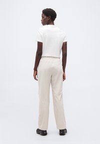 Άτομο που στέκεται με την πλάτη προς τα εμπρός, φοράει λευκό κοντομάνικο πουκάμισο, ανοιχτό μπεζ παντελόνι και μαύρα παπούτσια σε απλό λευκό φόντο.