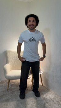 Άνδρας που χαμογελάει, στέκεται σε εσωτερικό χώρο φορώντας ένα λευκό γραφιστικό t-shirt, μαύρο παντελόνι και μαύρα παπούτσια, κοντά σε μια εκρού πολυθρόνα και ένα μικρό στρογγυλό τραπέζι.