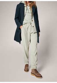 Tummansininen teksturoitu takki, vaaleanvihreä huppari, vaaleanvihreä ribbineuloksinen housut, ruskeat nauhalliset kengät paksulla pohjalla ja koristeellisilla yksityiskohdilla.