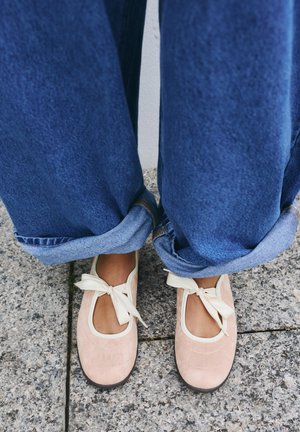 Personne portant un jean bleu retroussé et des ballerines rose pâle avec des nœuds en ruban crème debout sur un trottoir carrelé gris.