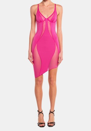 Vrouw draagt een doorschijnende, felroze, mouwloze mini-jurk met asymmetrische zoom en zwarte sandalen met hoge hakken, staand tegen een effen achtergrond.