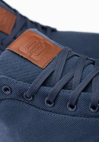 Zapatilla de lona azul marino con una etiqueta de cuero marrón, cordones redondos y ojetes. La tela tiene un acabado texturizado con detalles de costura visibles.