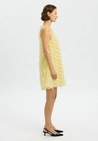 Yellow sleeveless dress with a textured, layered design. Features a loose fit and falls above the knees. Paired with black pointed slingback shoes.