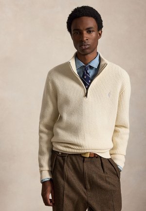 Jeune homme portant un pull en maille crème par-dessus une chemise bleue et une cravate à motifs, avec un pantalon marron à carreaux et les mains dans les poches.