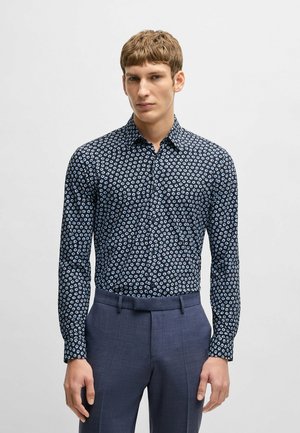 Camicia blu navy con un piccolo motivo floreale bianco, maniche lunghe e colletto. Abbinata a pantaloni grigi su misura.