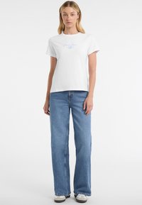 Camiseta blanca de algodón con mangas cortas y cuello redondo, con un logotipo en texto azul claro. Combinada con unos vaqueros de tiro alto de pierna ancha en azul claro.
