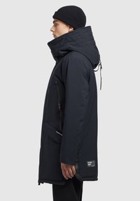 Parka de un albastru închis cu guler înalt, glugă ajustabilă, buzunare laterale și un material texturat. Prezintă accente negre și un ecuson cu logo.