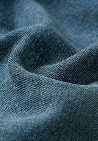Denim-kangasta syvässä sinisessä sävyssä, esiteltynä tekstiilien rakenteella, näkyvillä ompeleilla ja pehmeällä valuvalla olemuksella, heijastaen valoa hienovaraisesti.
