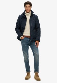 Marineblauwe jas met rits, met toggle-details en voorzakken, gecombineerd met een crèmekleurige coltrui, blauwe spijkerbroek en tan veterschoenen.