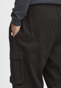 Sorte cargo-bukser med en sidelomme, lavet af slidstærkt stof. Har en afslappet pasform og elastisk talje. Lille label synlig på lommen.