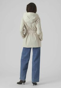 Manteau trench beige avec capuche, taille ceinture et poches latérales ; porté sur un jean large bleu clair et des bottines à talons noires.