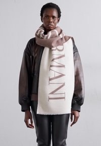 Veste aviateur en cuir marron, pantalon en cuir noir et une écharpe crème avec "ARMANI" en texte marron. L'écharpe a des bords à franges et une texture douce.
