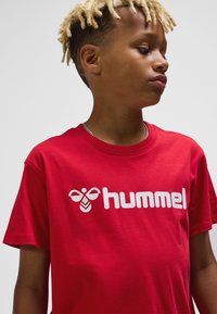 Röd bomulls-t-shirt med en framträdande vit "hummel"-logotyp och bi-motiv. Klassisk rund halsringning och korta ärmar med slät textur.