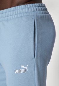 Svetlo modri športni pajac iz mehkega blaga, ki ima elastičen pas, stransko žepek in belo vezeno Puma logotip.