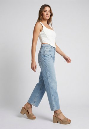 Vrouw in een witte mouwloze top, lichtblauwe jeans en beige plateauzandalen die tegen een effen grijze achtergrond loopt.