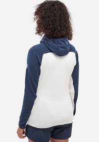 Person with curly hair wears a white and navy long-sleeve hooded jacket and navy shorts, shown from behind against a white background.