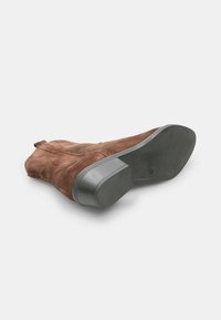 Bota de ante marrón hasta el tobillo con punta redonda, diseño minimalista, tacón plano y minimal de herrajes. La suela es texturada y ligeramente curvada.