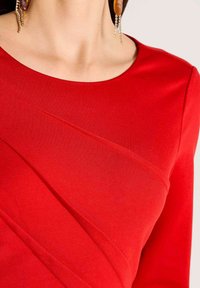 Rotes Langarmshirt mit rundem Ausschnitt und diagonalen Steppnähten als texturgebende Akzente. Glatter Stoff mit figurbetontem Schnitt.