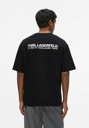 Man draagt een zwarte oversized T-shirt met witte tekst "Karl Lagerfeld 21 Rue St-Guillaume Paris" op de rug, gecombineerd met grijze pantalon met fijne strepen.