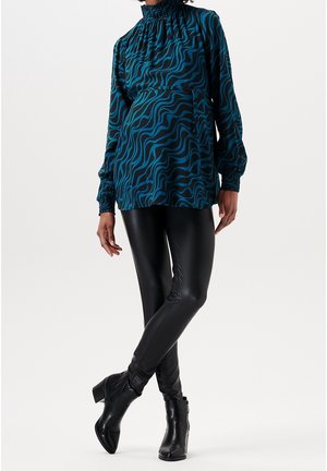 Femme portant un chemisier à motif ondulé bleu sarcelle et noir à manches longues, des leggings en cuir noir et des bottines noires sur un fond uni.