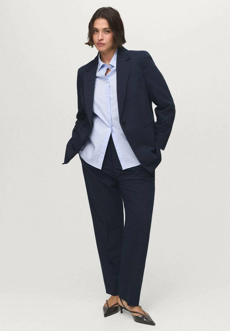 Donna in piedi con le mani in tasca, che indossa un completo blu navy, una camicia azzurro chiaro e scarpe nere con tacco basso a punta su sfondo neutro.