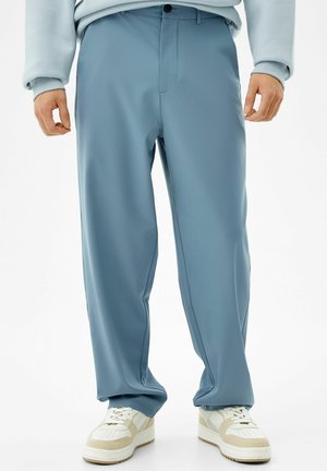 Homme portant un pantalon ample bleu clair, des baskets blanches et beiges, et un sweat léger, debout devant un fond blanc uni.