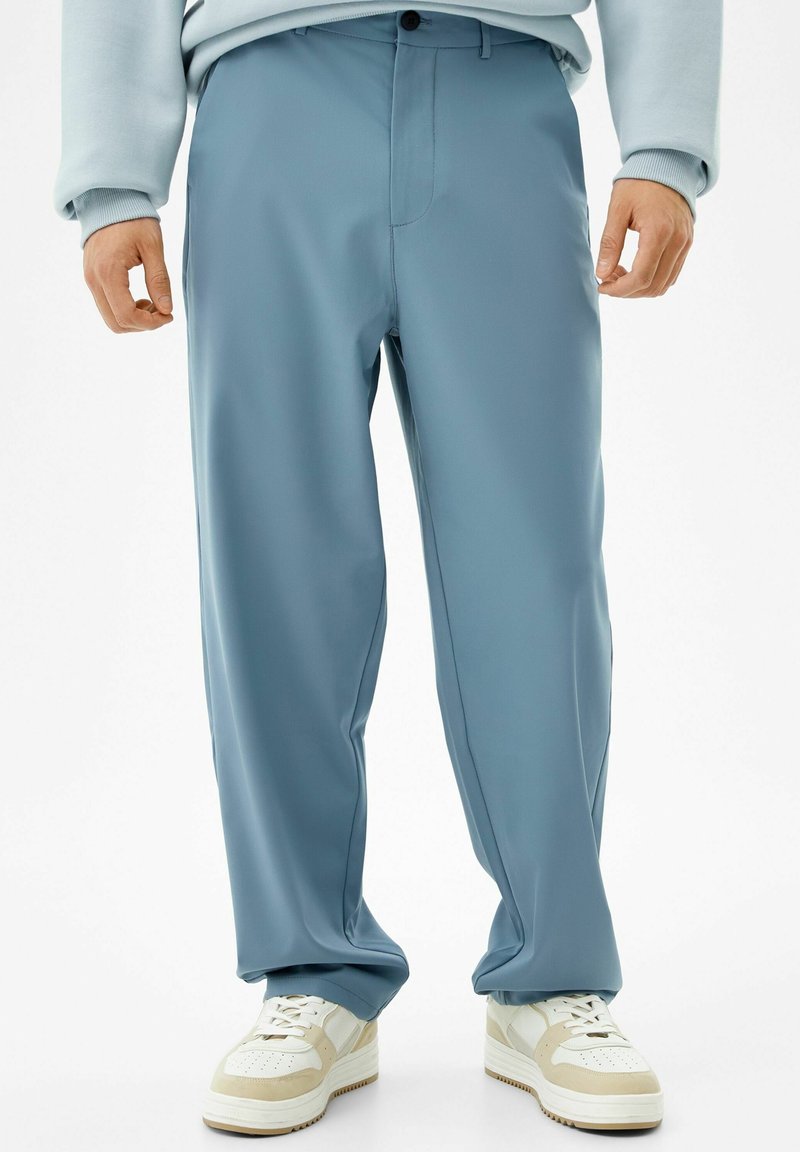 Homme portant un pantalon ample bleu clair, des baskets blanches et beiges, et un sweat léger, debout devant un fond blanc uni.