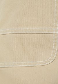 Beżowa tkanina o gładkiej fakturze, z białym przeszyciem i widocznym szwem wzdłuż krawędzi, wskazującym na szczegół konstrukcyjny odzieży.