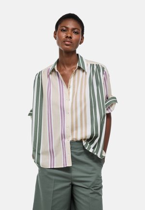 Femme debout, les mains dans les poches, portant une chemise boutonnée oversize à rayures verticales vertes, beiges et violettes, accompagnée d'un pantalon vert.