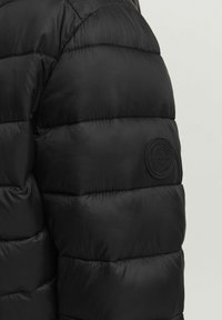 Zwarte gewatteerde pufferjack-sleeve met circulaire, reliëf geperste merkembleem, gladde glanzende stof en horizontale stikdetails.