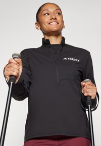 Pullover nero a maniche lunghe con zip a un quarto e tasca frontale, con logo TERREX. Utente tiene in mano bastoni da trekking con manici in gomma.