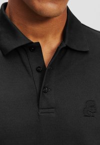 Tricou polo negru dintr-un material texturat, cu un guler cu două butoane și un emblema brodată pe partea stângă a pieptului.