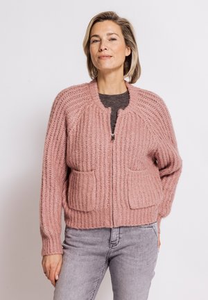 Woman wearing a pink chunky knit zip-up cardigan with front pockets and light gray jeans against a plain background.