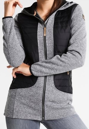 Mujer con chaqueta de forro polar gris con paneles acolchados negros, cremallera frontal, capucha y bolsillos con cremallera, posando con los brazos cruzados.