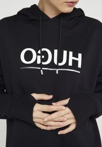 Person som bär en svart hoodie med vitt "HUGO"-logotyp, med händerna knäppta vid midjan,ser lite åt sidan.
