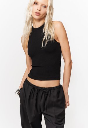 Blondynka z luźnymi falującymi włosami, ubrana w bezrękawnikowy czarny krótki top i luźne czarne spodnie ze sznurkiem, na białym tle.