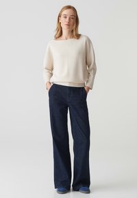 Maglione a coste crema con scollatura ampia, abbinato a jeans a gamba larga blu scuro e sneakers blu, in un contesto minimalista.