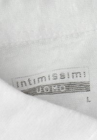 Bílá textilie s měkkou strukturou, na které je štítek s nápisem "intimissimi", velikost "L", šedým písmem. Tkanina vypadá mírně pleteně.