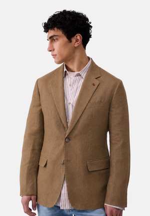 Giovane uomo con capelli corti e ricci che indossa un blazer marrone sopra una camicia a righe con bottoni, guarda verso sinistra su uno sfondo semplice.
