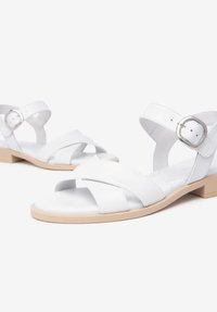 Sandales en cuir blanc avec des brides croisées et une sangle à la cheville avec boucle. Semelle beige plate avec un léger talon. Texture lisse, design minimaliste.