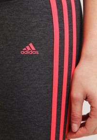 Leggings de sport grises Adidas avec trois rayures verticales roses et un petit logo rose. Gros plan sur la texture du tissu et les détails du design.