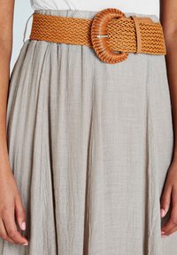 Vävt tanfärgat bälte med en matchande cirkulär spänne, med en texturerad design, bärs över en ljusgrå kjol.