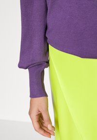 Fialový pletený svetr s ribbovanými manžetami, spárovaný s jasně žlutou sukní s diagonálním švem. Hladká textura a živé barvy.