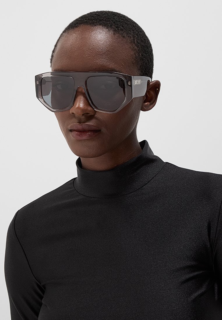 Lunettes de soleil surdimensionnées avec une monture transparente gris foncé et des verres foncés. Surface lisse avec des bords anguleux. Haut à col roulé noir porté en dessous.