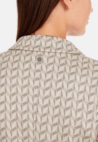 Beige patterned blazer with geometric design, textured fabric, notched collar, and small metallic logo accent on the back.