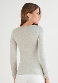 Světle šedý top s dlouhým rukávem vyrobený z měkké látky. Má krajkový lem na zápěstí, kulatý výstřih, přiléhavý střih a hladkou strukturu.