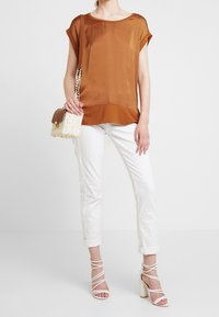 Top de seda marrón, de manga corta y con dobladillo curvado, combinado con pantalones blancos enrollados y tacones blancos con tiras. Lleva un bolso de mimbre.
