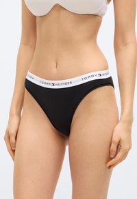 Γυναίκα που φοράει μαύρο εσώρουχο σε στυλ μπικίνι Tommy Hilfiger με λευκή ζώνη μέσης με το λογότυπο και λευκό σουτιέν, στέκεται μπροστά σε ένα απλό φόντο.