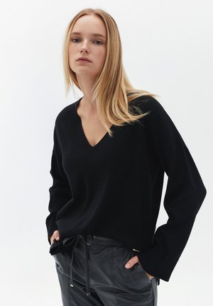 Pull noir en tricot avec un col en V, doté de poignets côtelés et d'une coupe décontractée. Porté sur un pantalon noir à taille élastique.