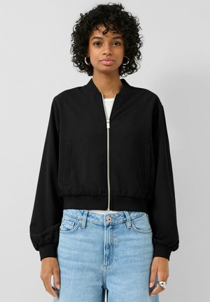 Kobieta ubrana w czarną bomberkę zapinaną na zamek, na białej bluzce i jasnoniebieskich dżinsach z wysokim stanem, stoi na tle jednolitego jasnoszarego tła.